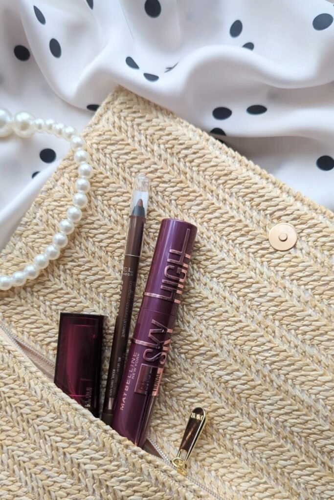 Burgundy mascara, brown eyeliner, and lipstick peeking from a woven straw clutch beside pearls and polka-dot fabric, styled for effortless beauty on the go.